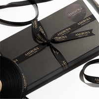 A black gift voucher package with the brand name 'Amirova Cosmetic Clinic' displayed prominently. The package is tied with a black ribbon that has the brand name printed on it.